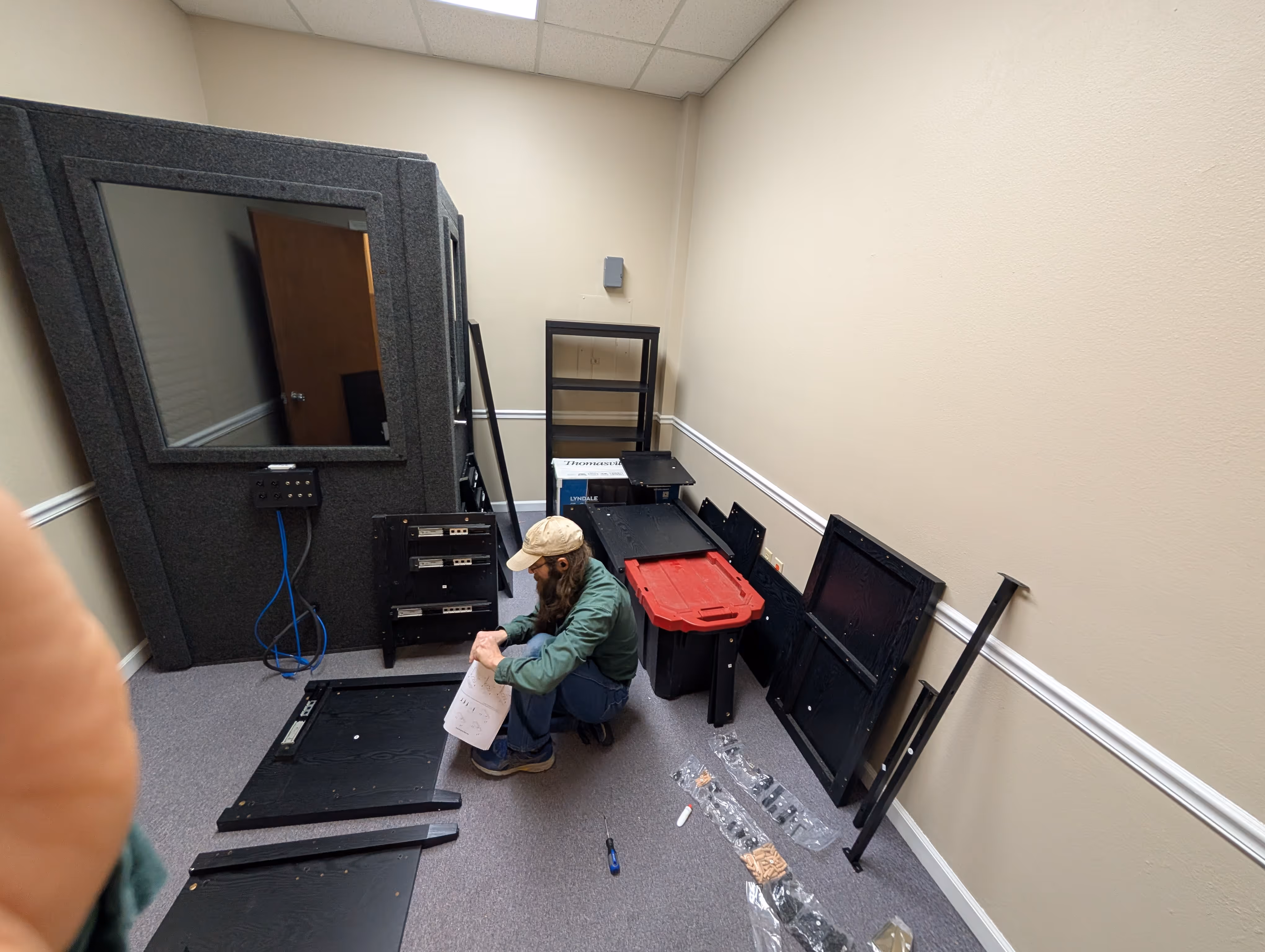 Man wearing a beige cap and green jacket assembling black wooden furniture pieces in a small room with beige walls and carpeted floor.