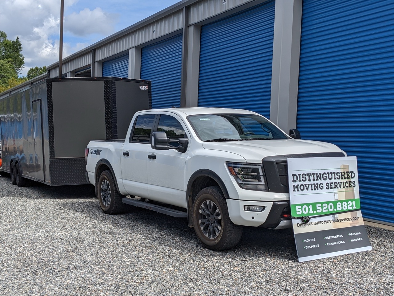 White pickup truck parked in front of blue storage unit doors with a trailer and a sign advertising Distinguished Moving Services and phone number 501.520.8821.