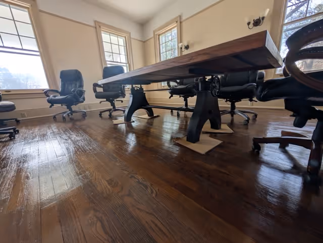 Conference room with a long wooden table supported by black metal legs on cardboard pieces and surrounded by black office chairs on a polished hardwood floor.