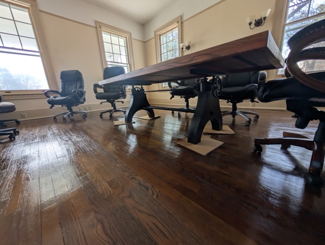 Conference room with a long wooden table supported by black metal legs on cardboard pieces and surrounded by black office chairs on a polished hardwood floor.