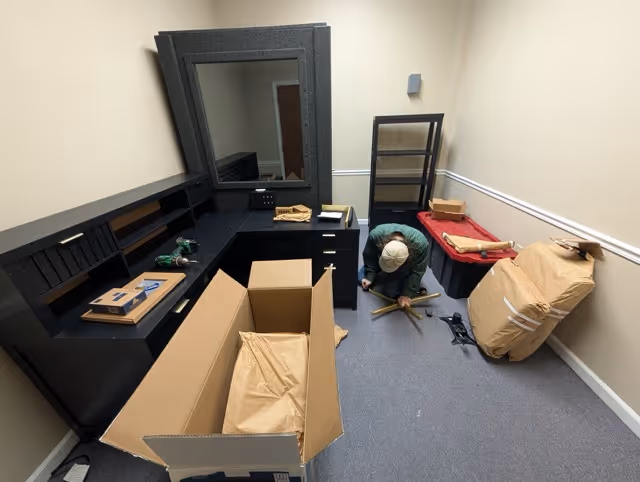 Person assembling or disassembling office furniture in a small room with packing boxes, wrapped chair, and black shelves.