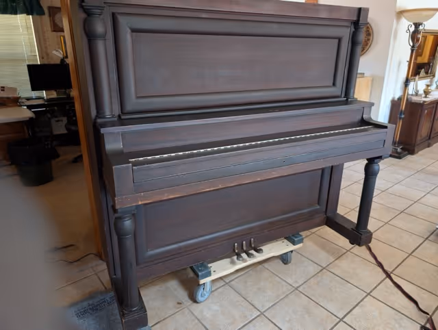 Dark wooden upright piano on a small dolly with wheels in a tiled room.