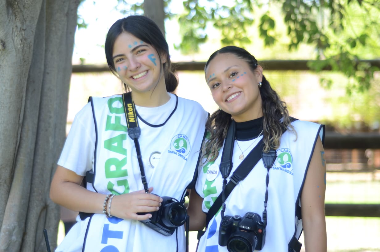 Dos jóvenes sonrientes con maquillaje azul en la cara, vistiendo chalecos blancos con el logo de Camp Santa Ursula y cámaras colgadas al cuello, posan juntas al aire libre con árboles y un cercado de madera al fondo.