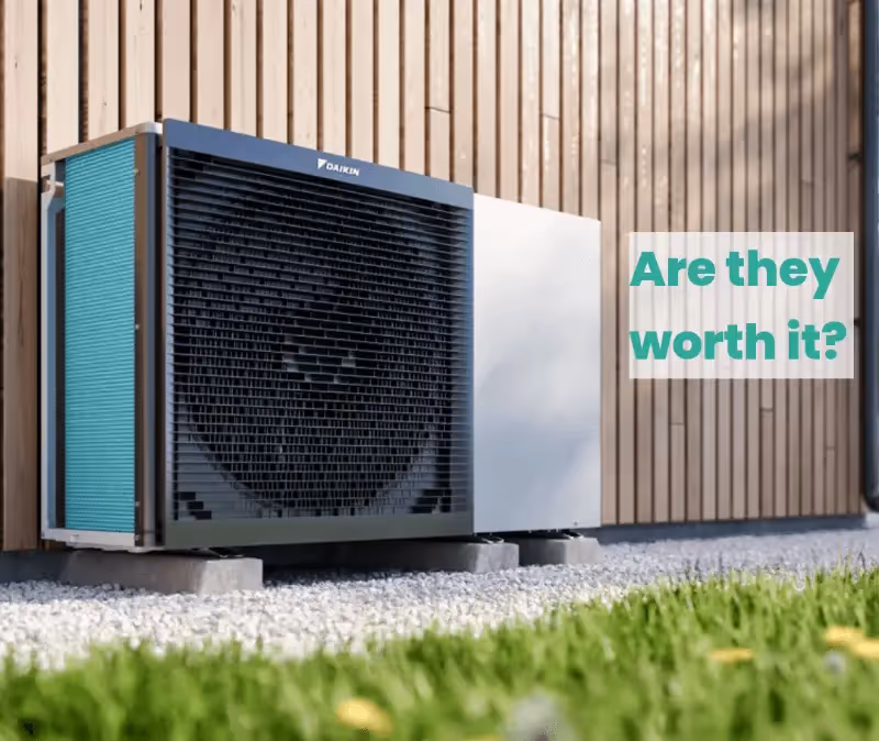 A heat pump on a path by a brown wall, with the text 'Are they worth it?' written above it