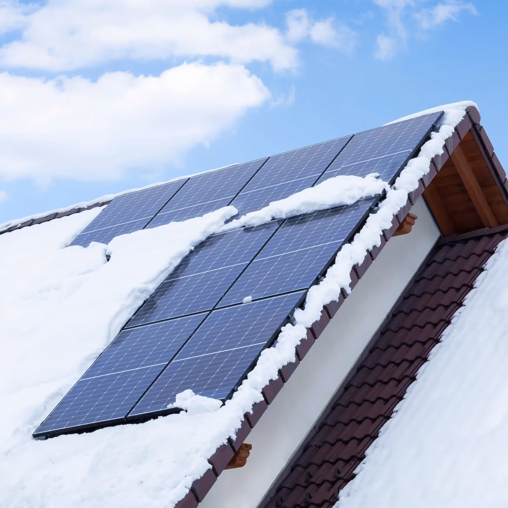 An image of a house with snow on the roof, solar panels sticking our from the snow and a clear blue sky