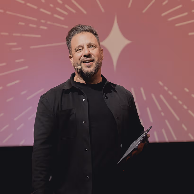 Man with short hair and beard speaking on stage, holding a tablet, with a pink background featuring a starburst design.