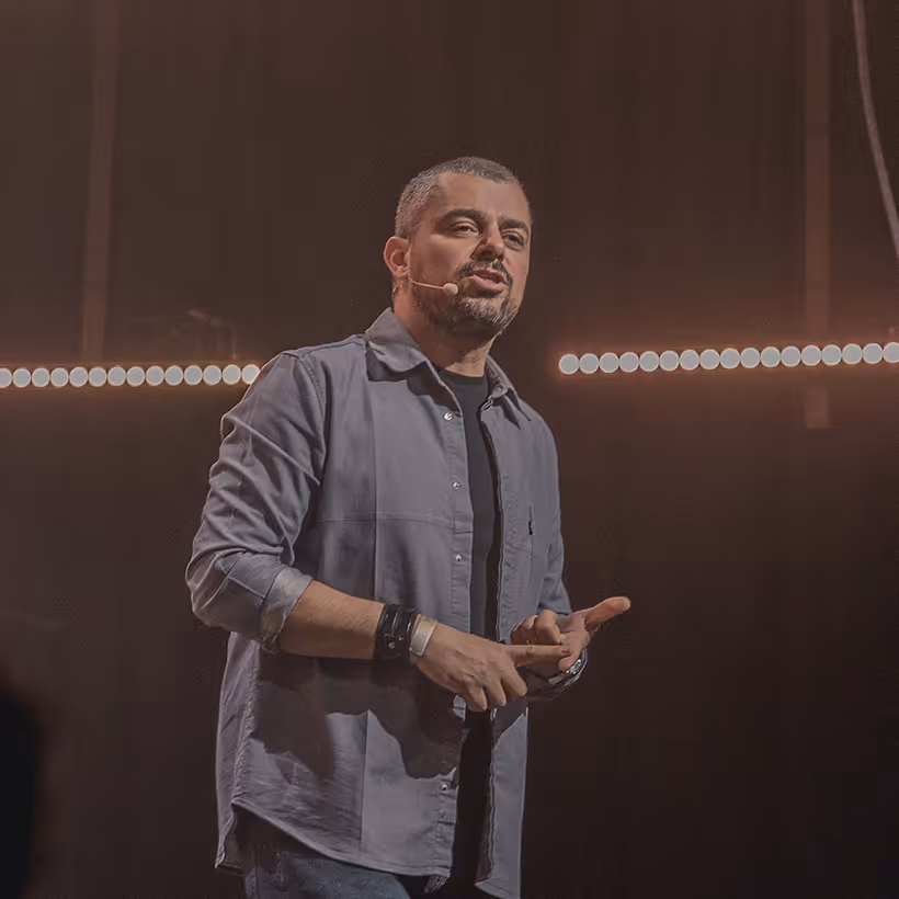Man with short hair and beard speaking on stage wearing a microphone headset and a gray button-up shirt.