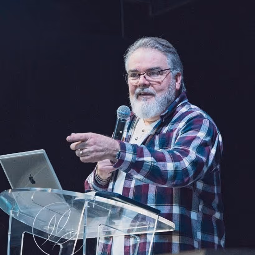Bearded man wearing glasses and a plaid shirt speaking into a microphone behind a transparent podium with a laptop.