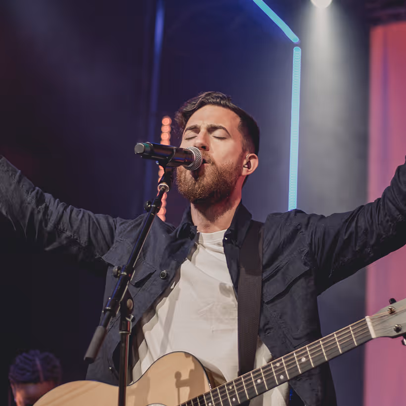 Bearded man singing passionately into a microphone while playing an acoustic guitar on stage with arms raised.