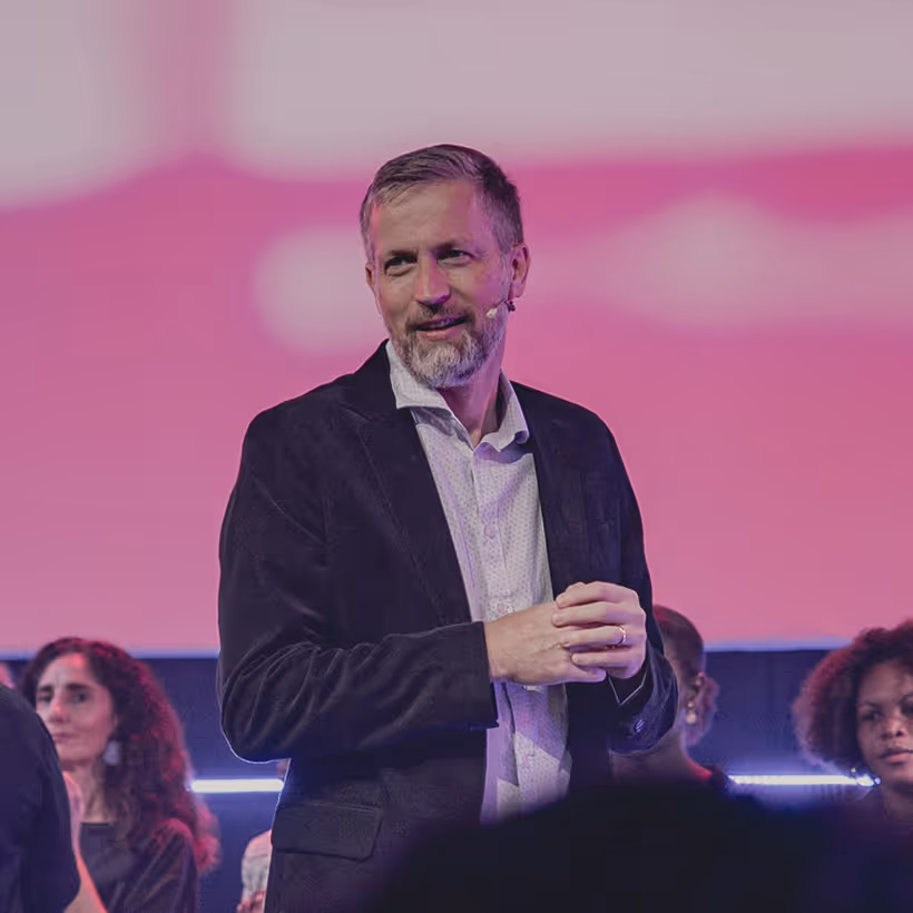 Man with a beard and microphone speaking on stage with people in the background under pink lighting.