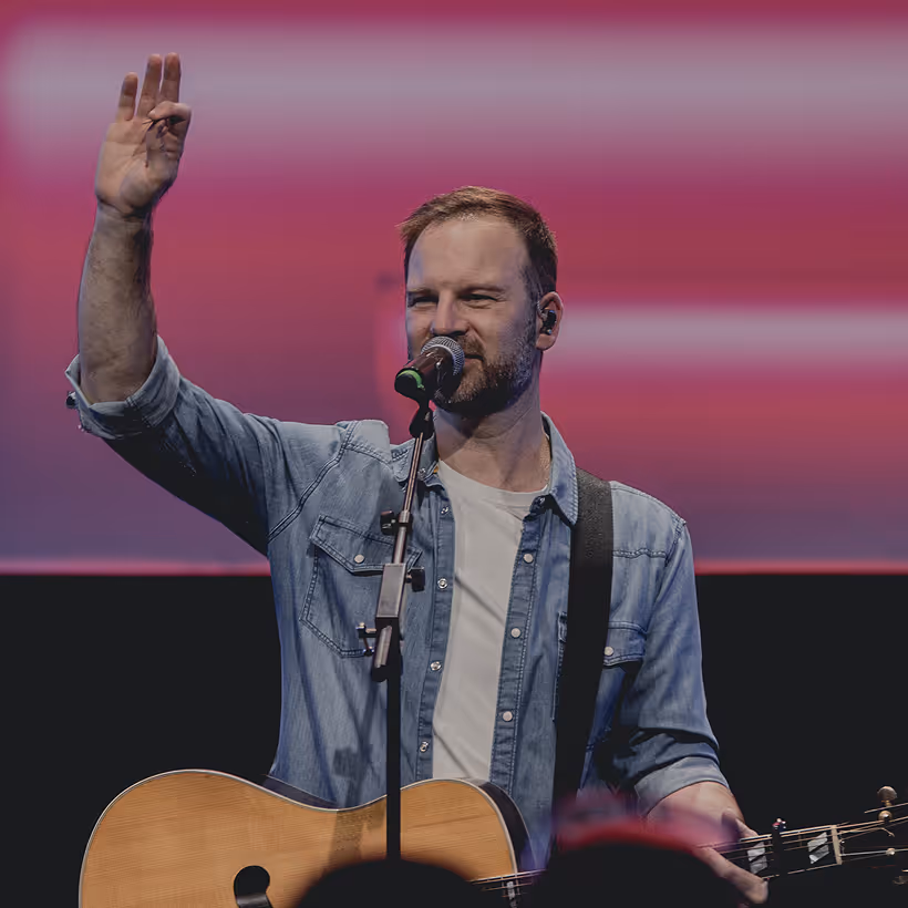 Male musician playing guitar and singing into a microphone, raising his left hand with three fingers extended.