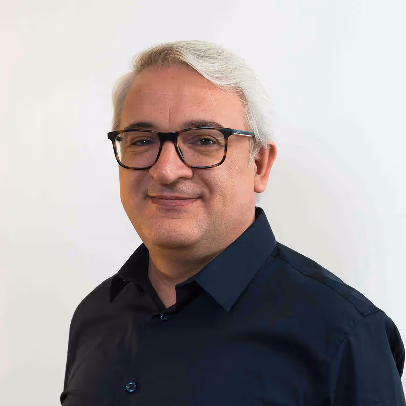 Smiling middle-aged man with white hair and black glasses wearing a dark blue shirt against a plain light background.