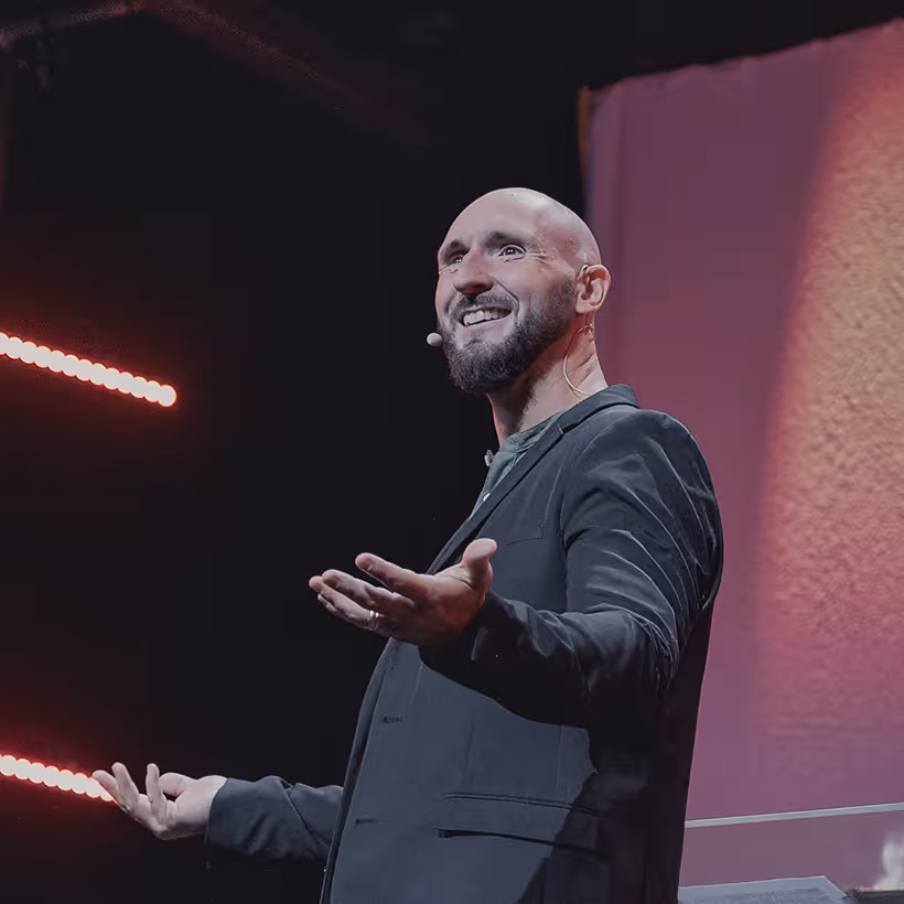 Smiling bald man with a beard speaking on stage wearing a black blazer and headset microphone.