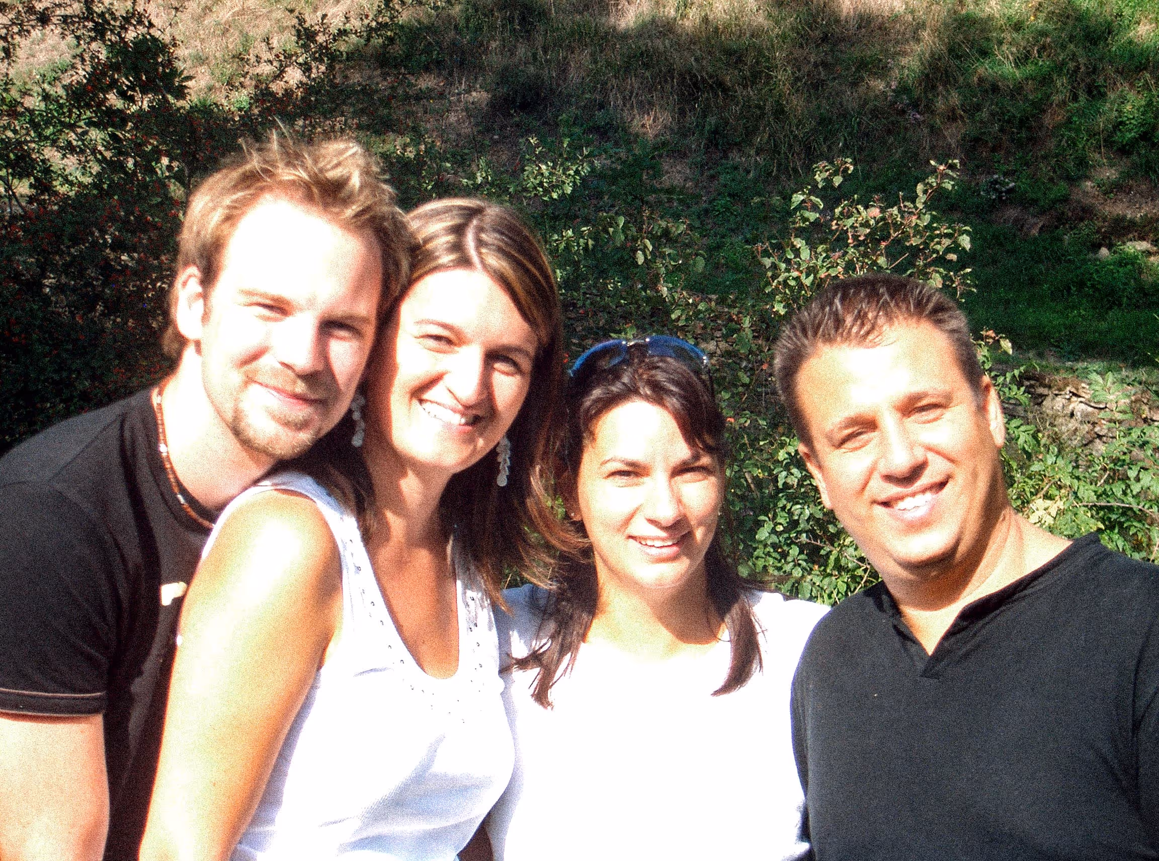 Four people smiling outdoors with greenery in the background on a sunny day.