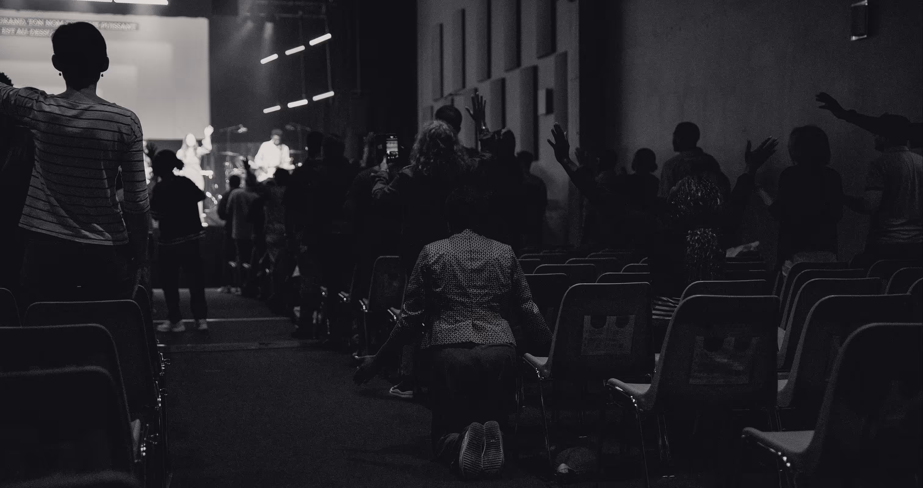 People in a darkened auditorium worshiping with raised hands; one person kneels in an aisle facing a lit stage.