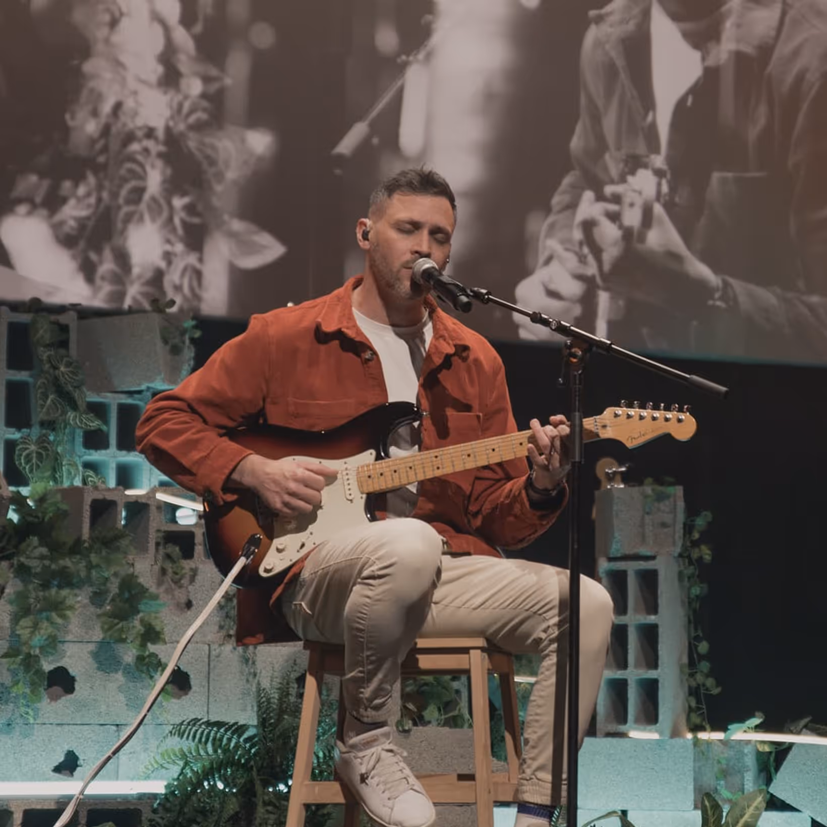 Man seated on a stool playing an electric guitar and singing into a microphone on a decorated stage.