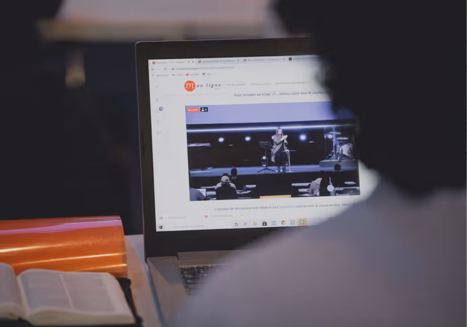 Person watching a live-streamed speaker on a laptop with an open book beside it.