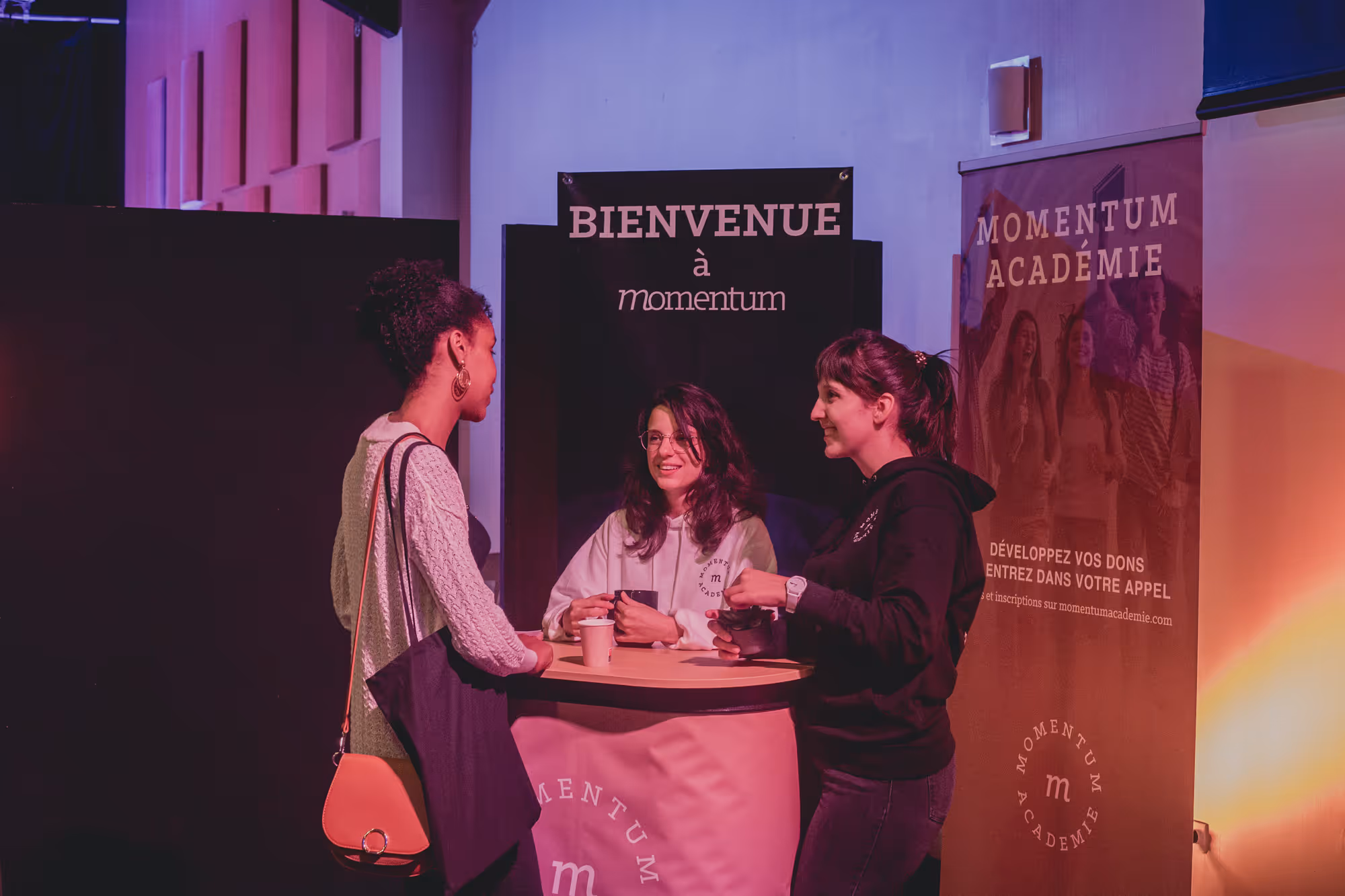 Three women conversing at a Momentum Académie booth with welcome signs in French.