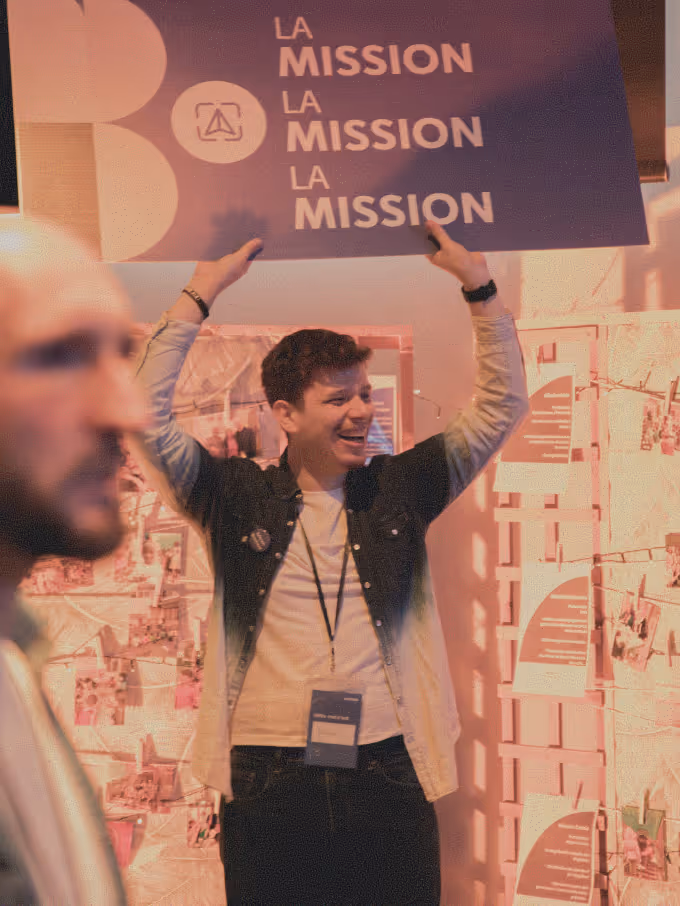 Smiling man holding up a blue sign that reads 'LA MISSION' three times in white letters at an indoor event.