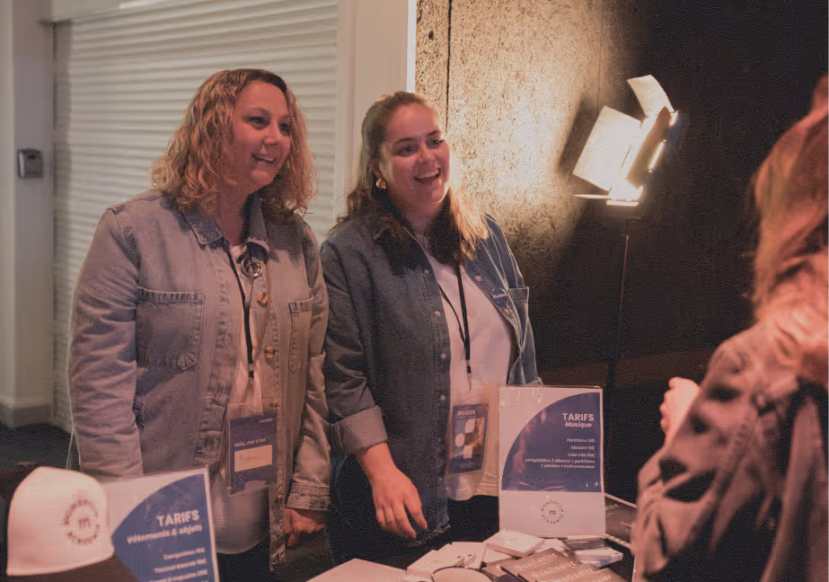 Two women in denim jackets smiling and talking to a person over a table with promotional materials and signs displaying prices.