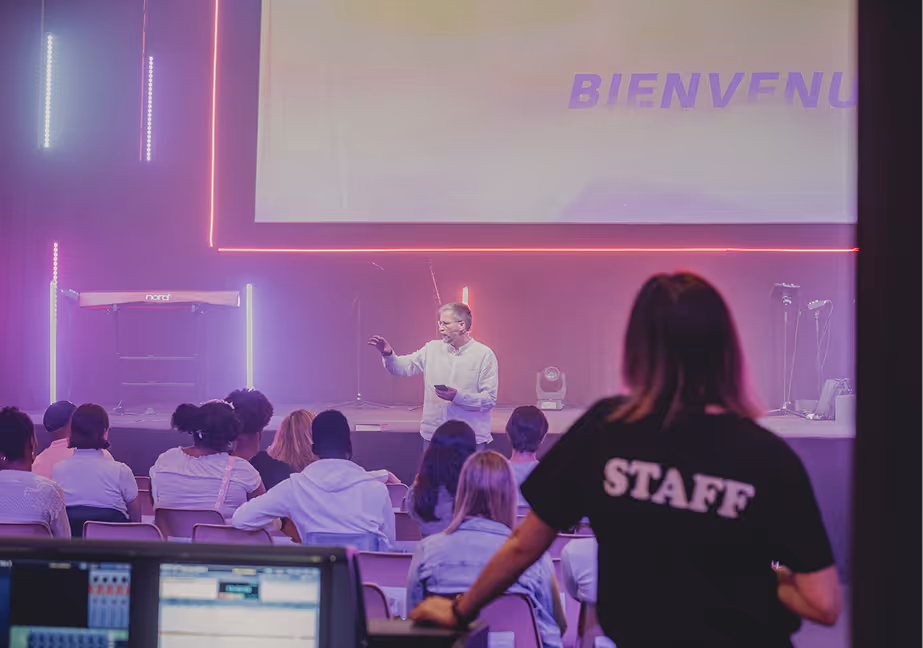 Speaker addressing an audience in a purple-lit auditorium with a staff member monitoring audio equipment.