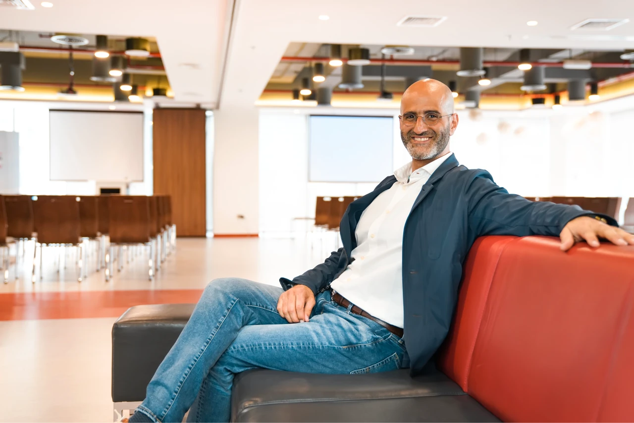 Smiling bald man with glasses in casual business attire sitting on a black and red couch in a modern conference room.