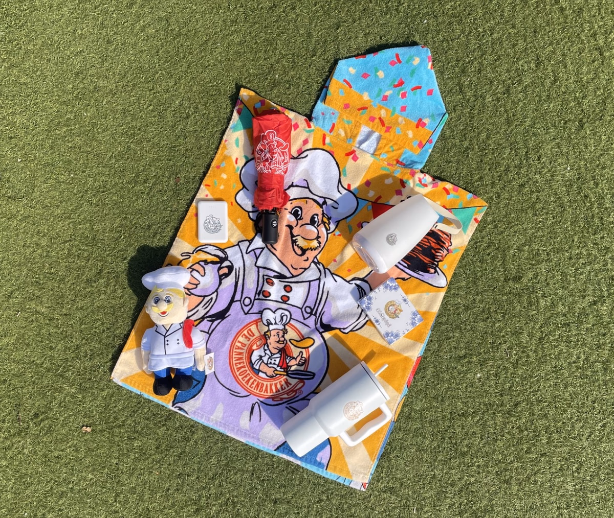 Verschillende voorwerpen waaronder een knuffel en thermosbeker in Pannekoekenbakker-thema op een badhandoek op een grasveld.