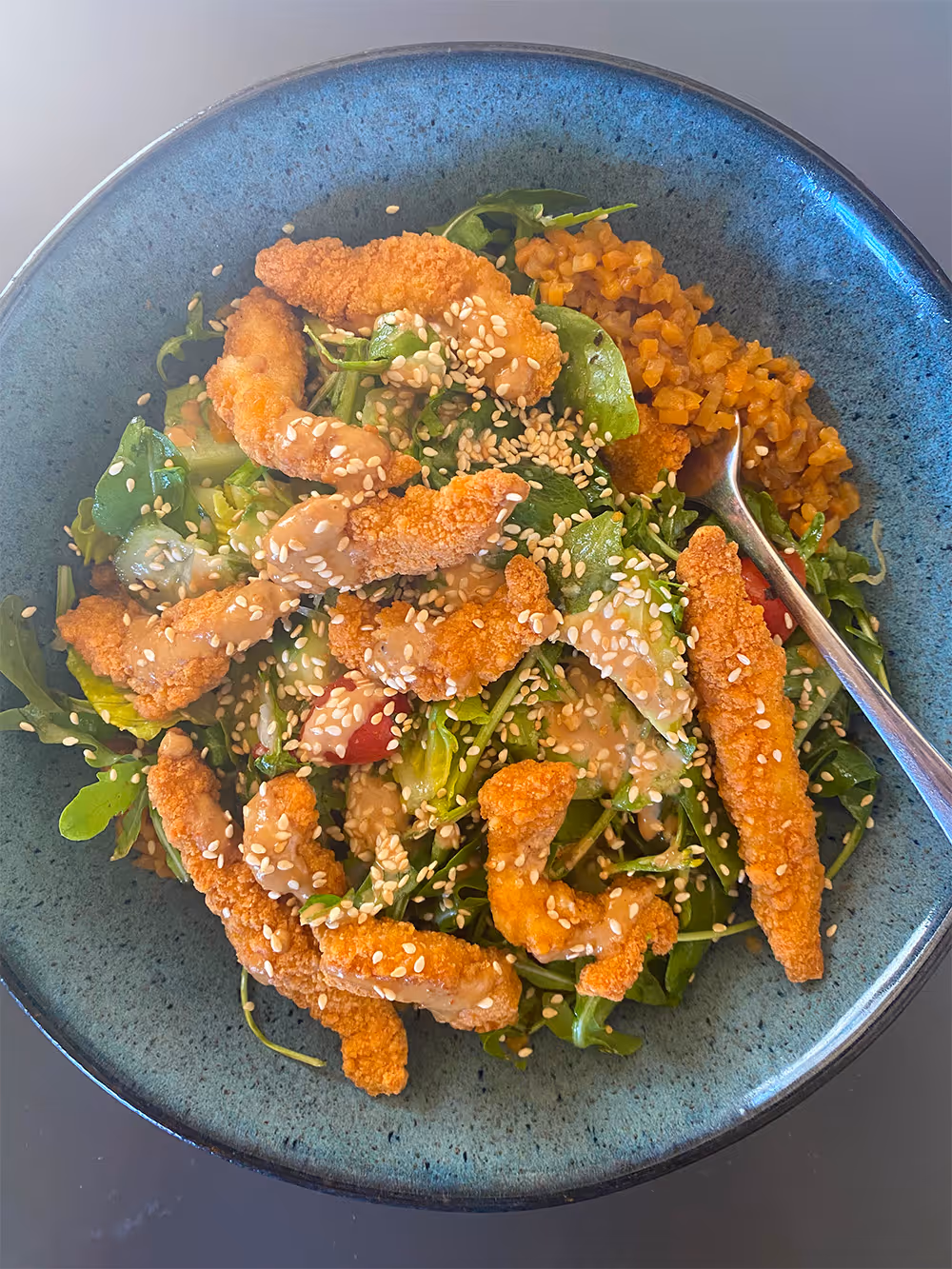 Salad bowl with mixed greens, cherry tomatoes, crispy breaded Shredded Chicken, sesame seeds, dressing, and a side of minced vegetables with a spoon.