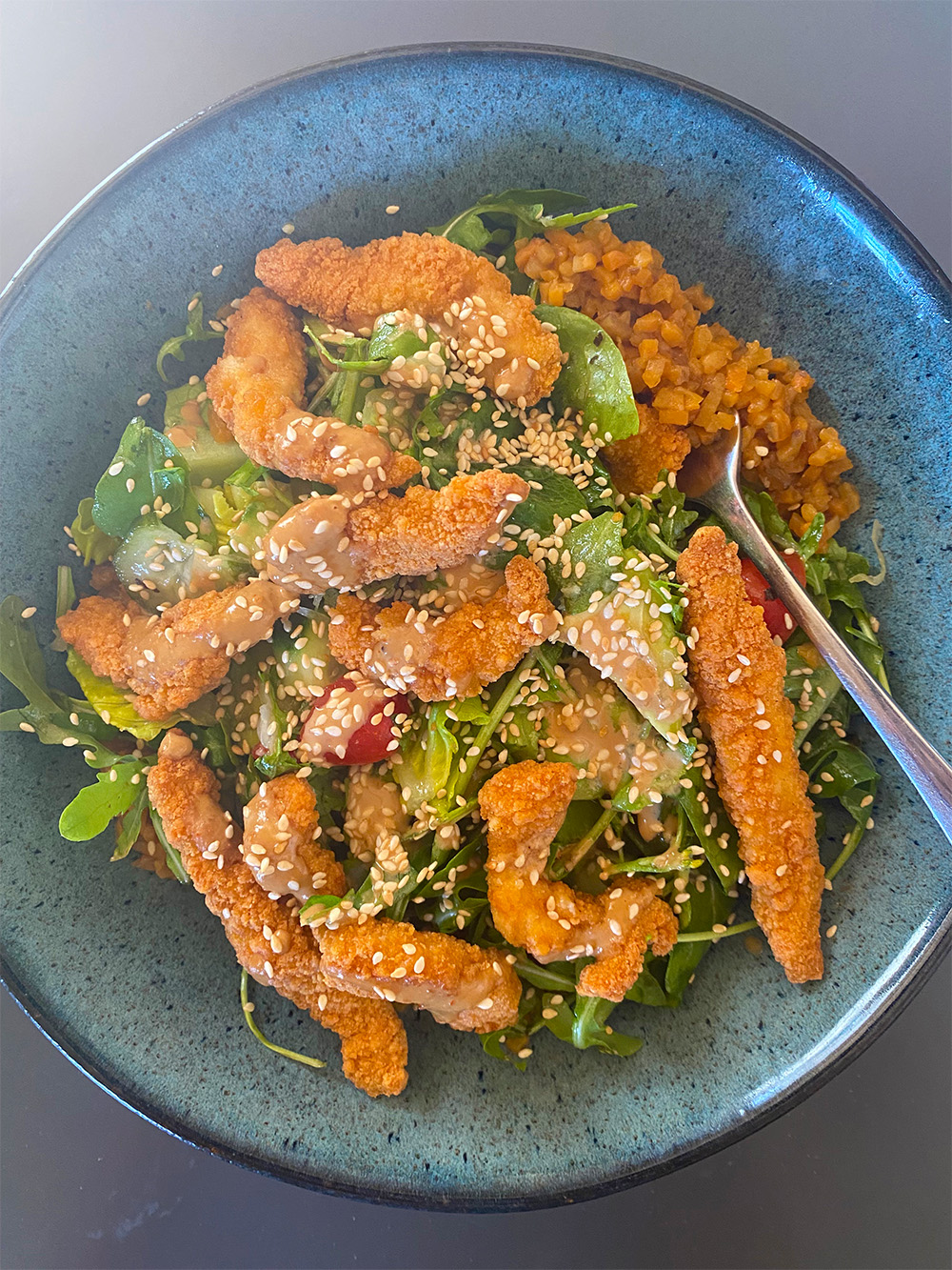 Salad bowl with mixed greens, cherry tomatoes, crispy breaded Shredded Chicken, sesame seeds, dressing, and a side of minced vegetables with a spoon.