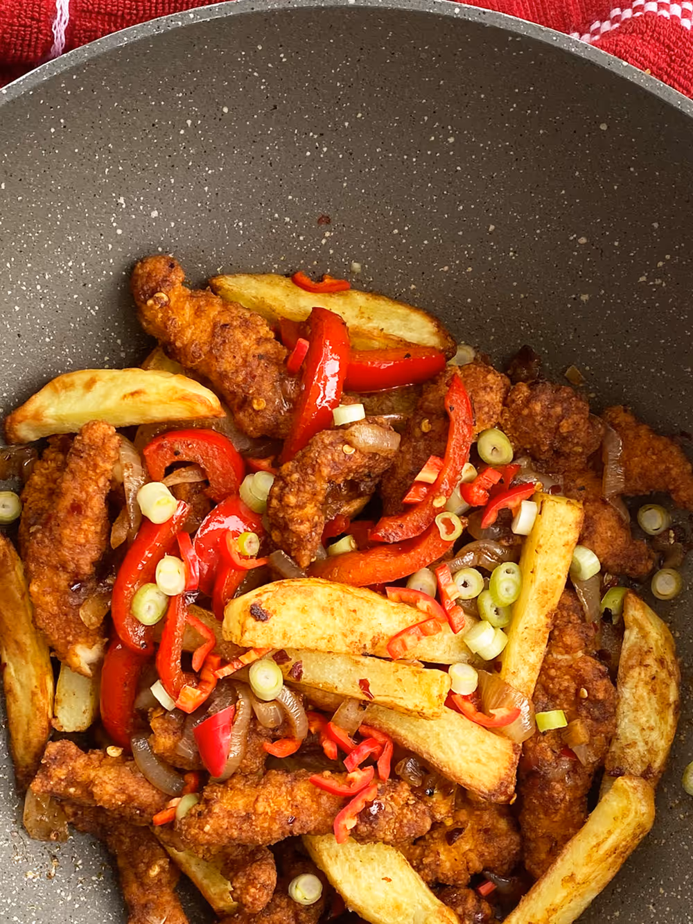Close-up of fried Shredded chicken, potato wedges, red bell peppers, sliced spring onions, and chili peppers in a pan.