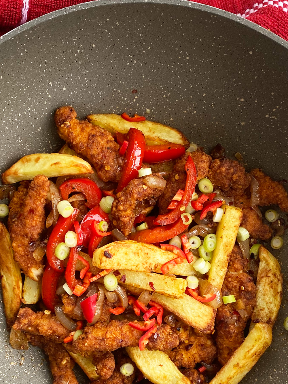 Close-up of fried Shredded chicken, potato wedges, red bell peppers, sliced spring onions, and chili peppers in a pan.