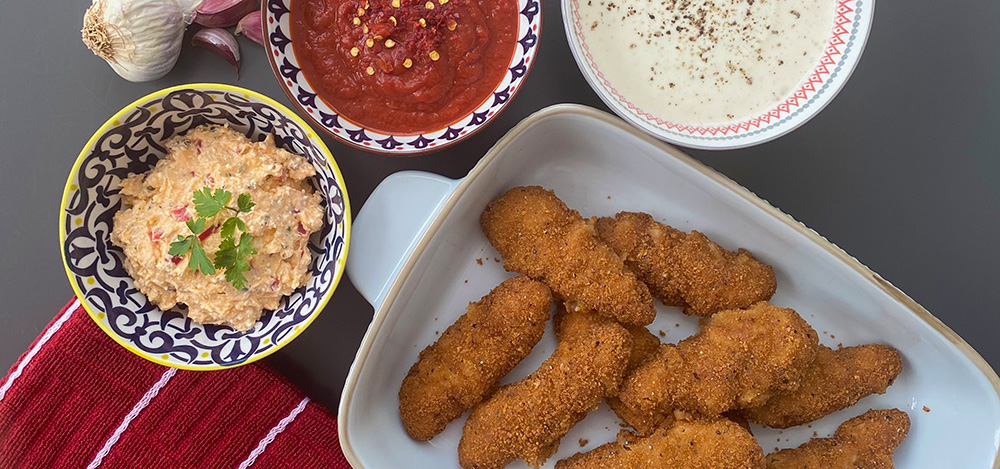 Plate of breaded chicken goujons with three bowls of dipping sauces and a garlic bulb on the side.