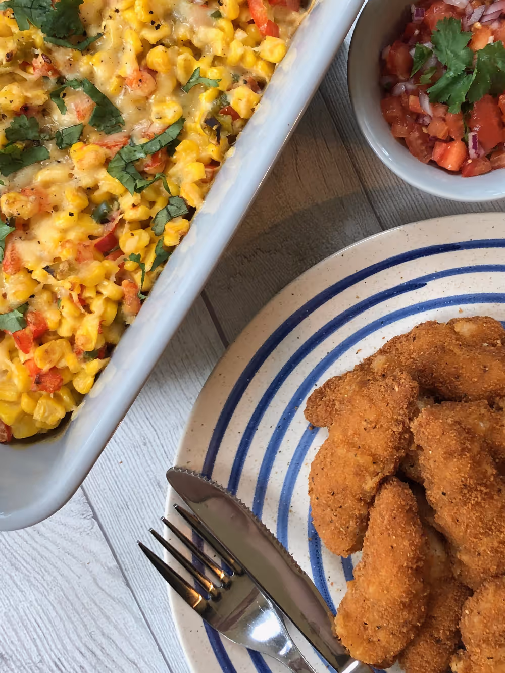 Plate with breaded chicken goujons, a dish of corn casserole topped with cheese and herbs, and a small bowl of tomato salsa with coriander.