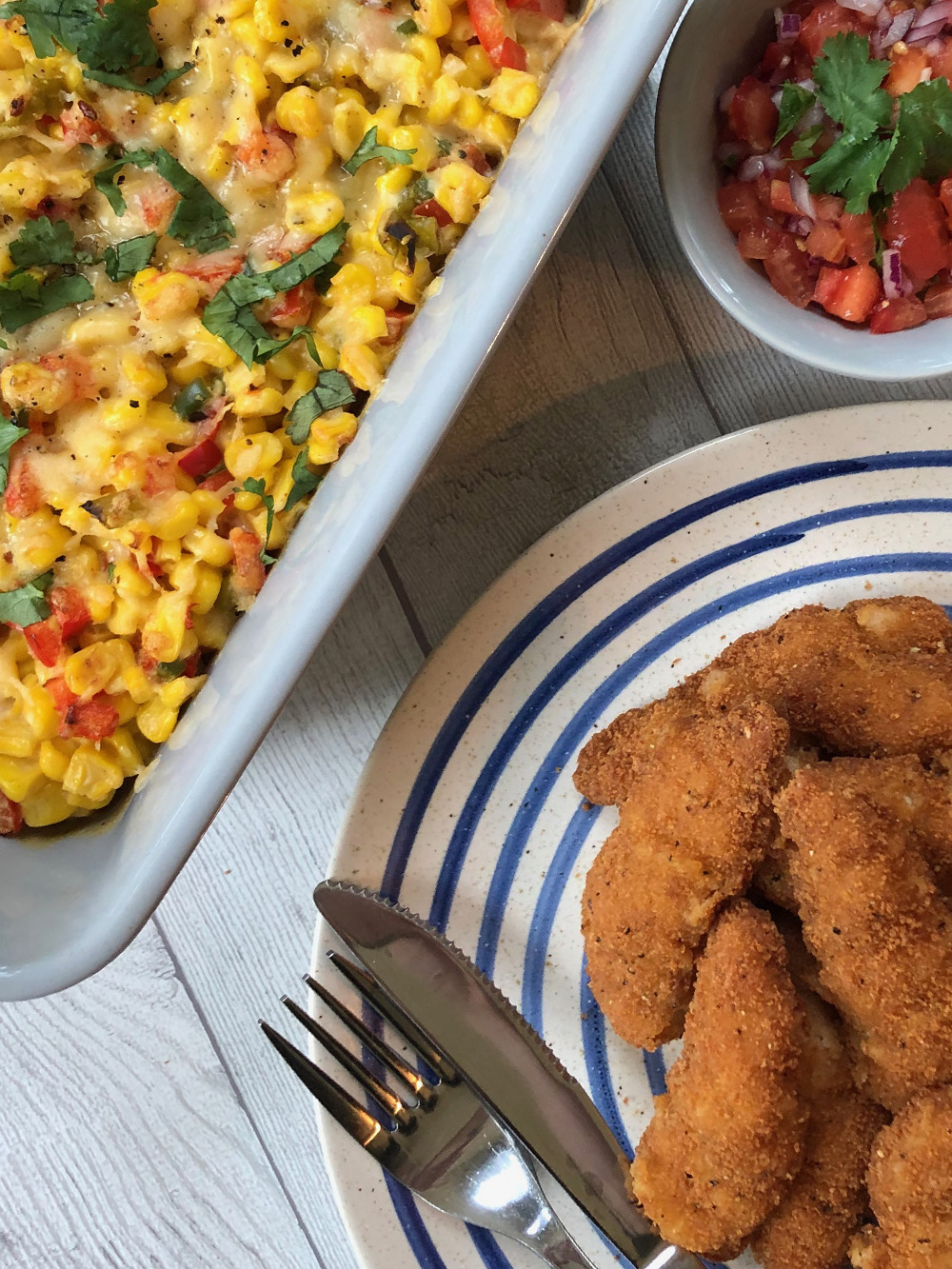 Plate with breaded chicken goujons, a dish of corn casserole topped with cheese and herbs, and a small bowl of tomato salsa with coriander.