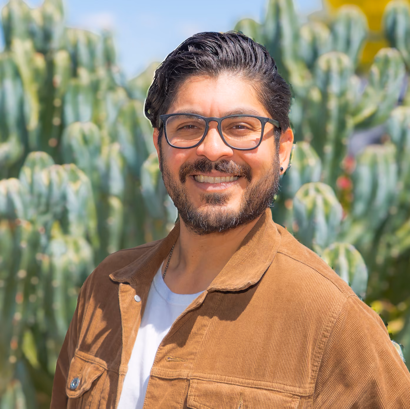 A portrait of Manny Diaz, a Latino man who is standing in front of a group of cacti. He has a smiling grin on his face and he is wearing a brown shirt jacket over a white t-shirt.