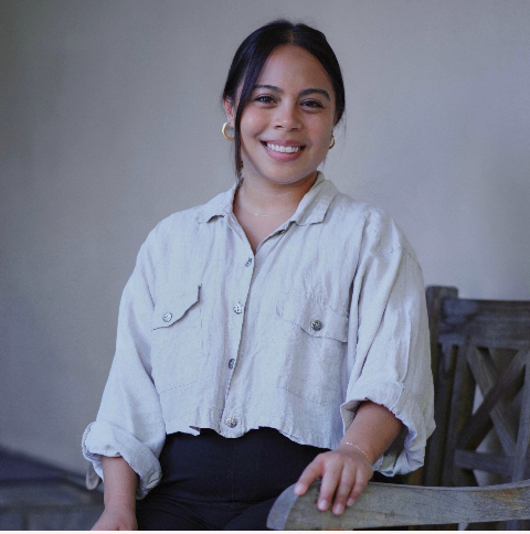 A portrait of Angela Medrano, a queer Latina person with long black hair that is mostly pulled back. She is wearing a gray button down and black pants, sitting in front of a gray background.