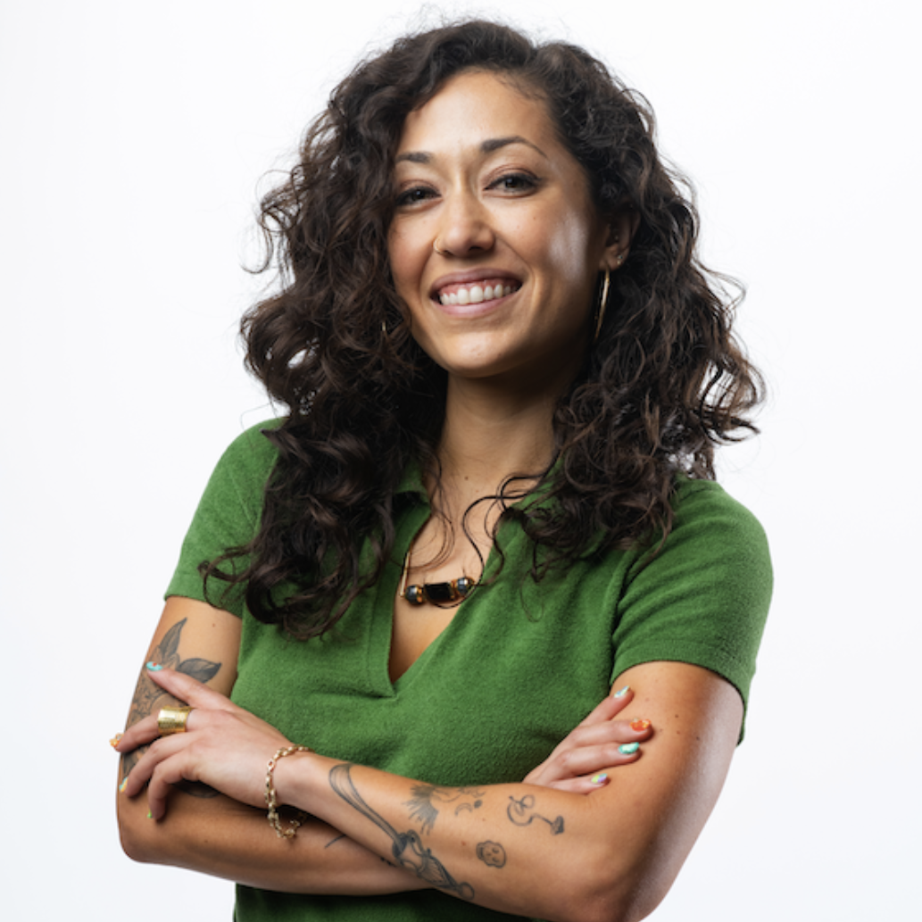 A portrait of Talia DeRogatis, a Latine woman with long, wavy black hair. She is standing in front of a white background with her arms crossed and a happy expression on her face. She is wearing a green v-neck shirt.