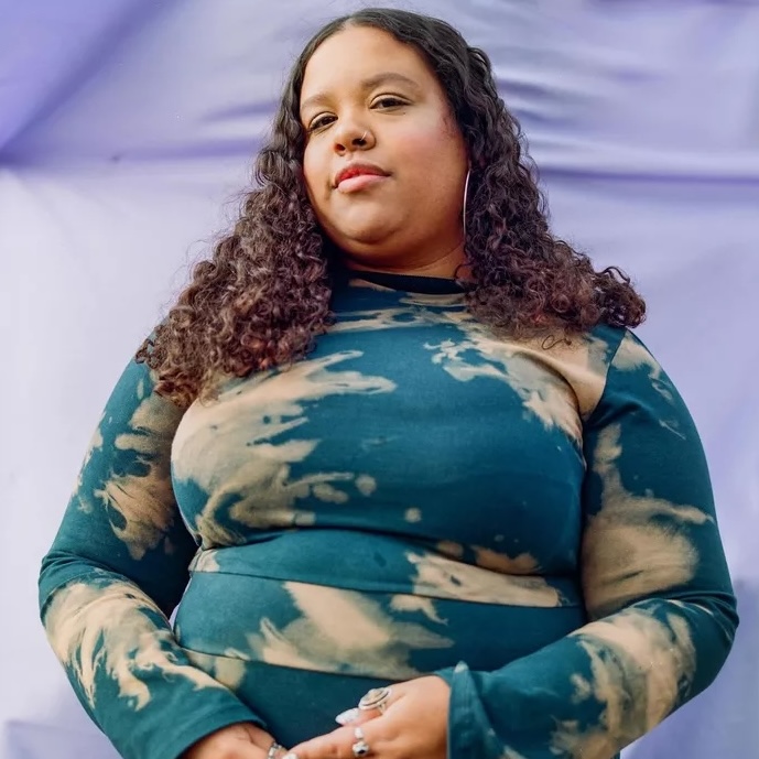 A portrait of Ashley M. Lagrange, a Black person with long black curls, hoop earrings, and a tie-dye shirt, standing in front of a lavender backdrop.