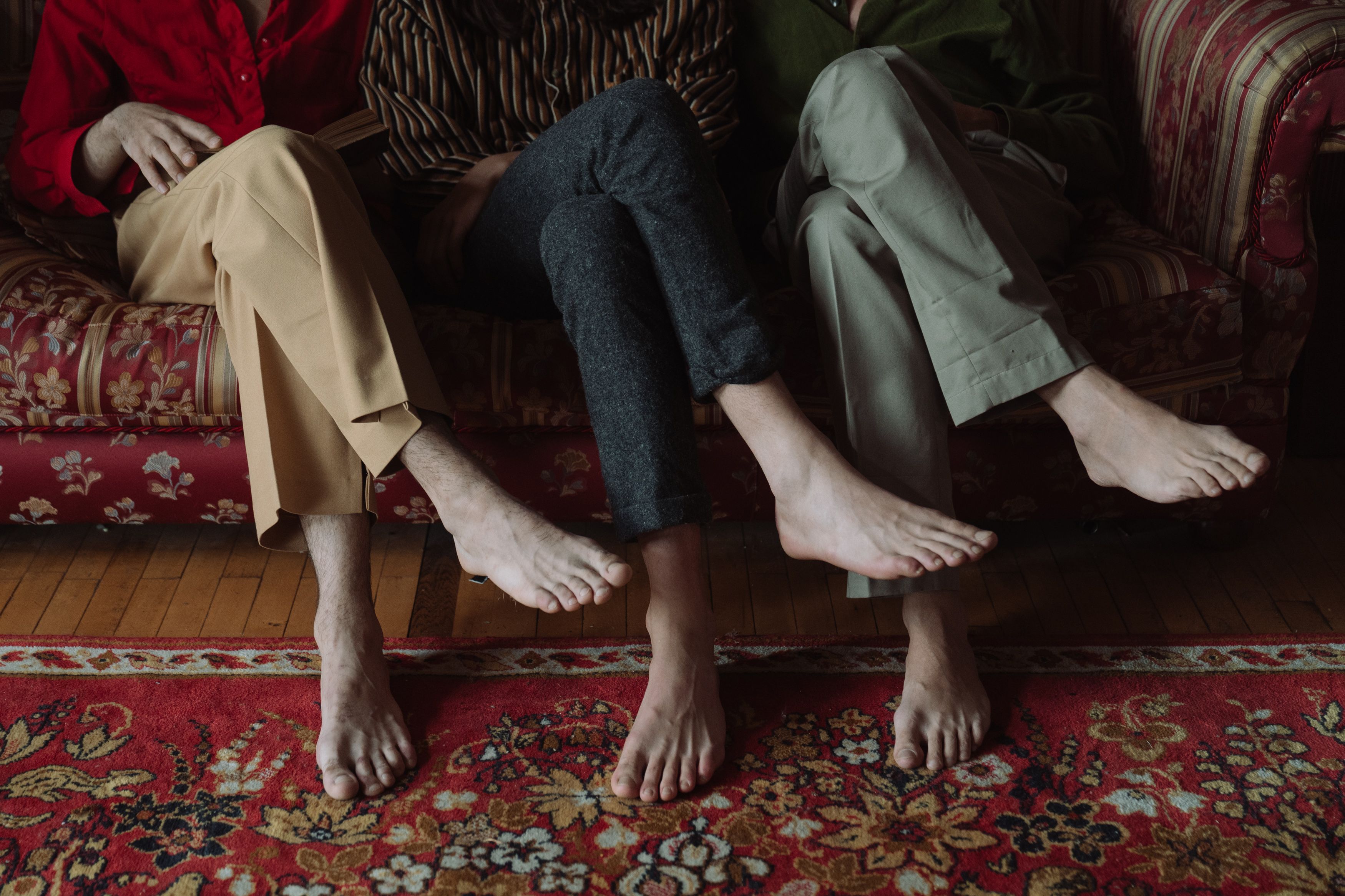 Three people sitting side by side on a vintage red sofa, bare feet crossed above a patterned rug.