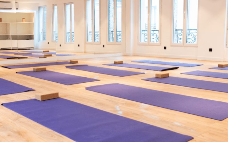 Salle de yoga lumineuse avec plusieurs tapis violets et blocs en bois sur un sol en bois clair.