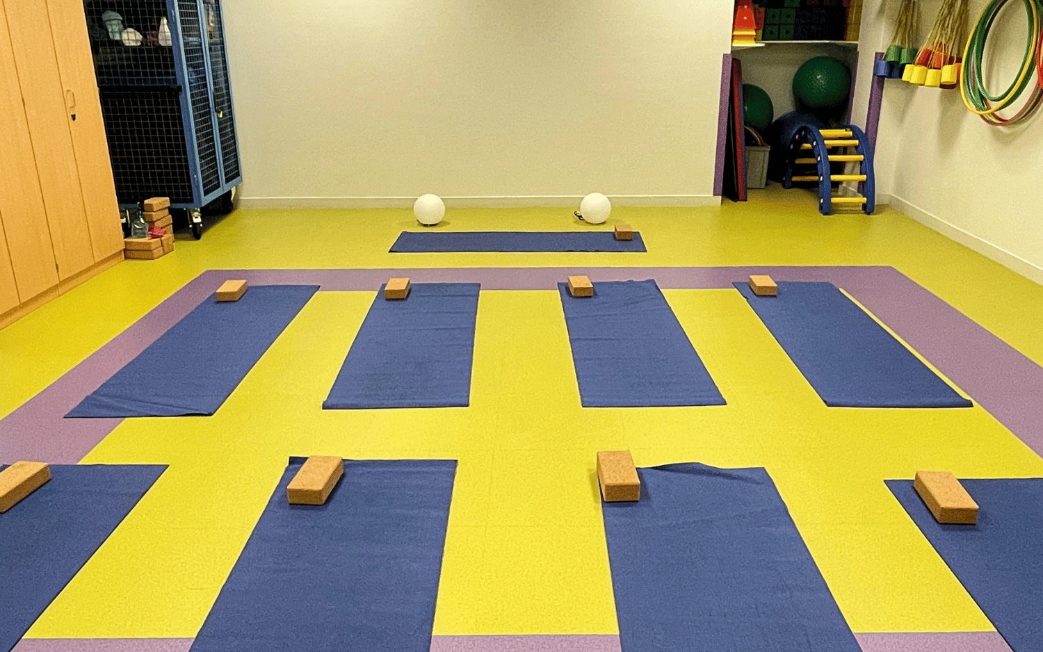 Salle de yoga avec plusieurs tapis bleus alignés sur un sol jaune et violet, chaque tapis équipé d'un bloc de yoga en bois.