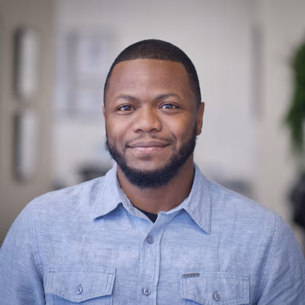 Smiling man with short hair and beard wearing a light blue button-up shirt in a blurred indoor setting.