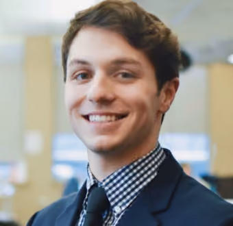 Young man smiling, wearing checked shirt, black tie, and dark blazer in an indoor setting.