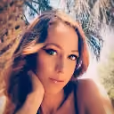 Portrait of a woman with long hair resting her face on her hand under palm tree fronds.
