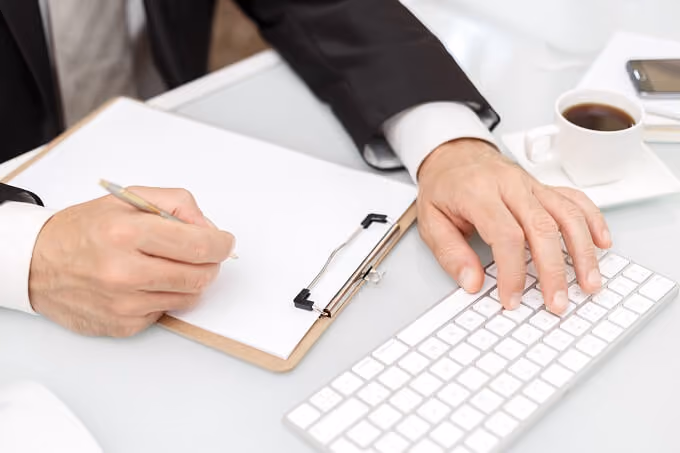 Person in suit writing on clipboard with one hand and typing on keyboard with the other at desk.