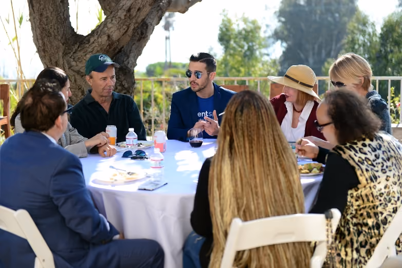 Group of eight people seated around a white round table outdoors, engaged in conversation and eating.