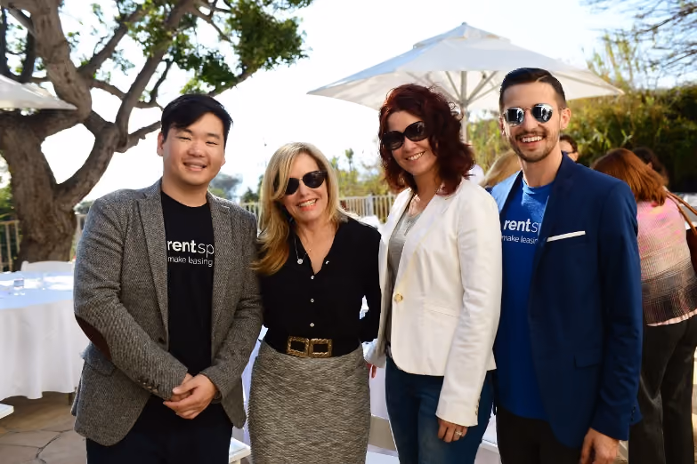 Four adults wearing sunglasses smile outdoors at an event under white umbrellas with trees in background.