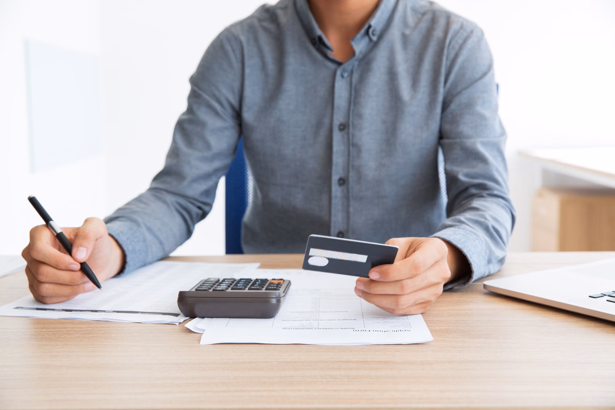 Person calculating expenses holding credit card and pen over documents with calculator on desk.