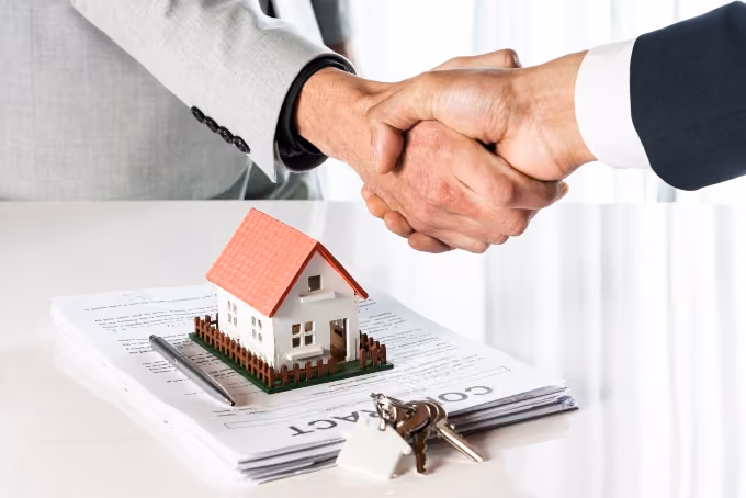 Two people shaking hands over a model house, a contract, and keys on a table.