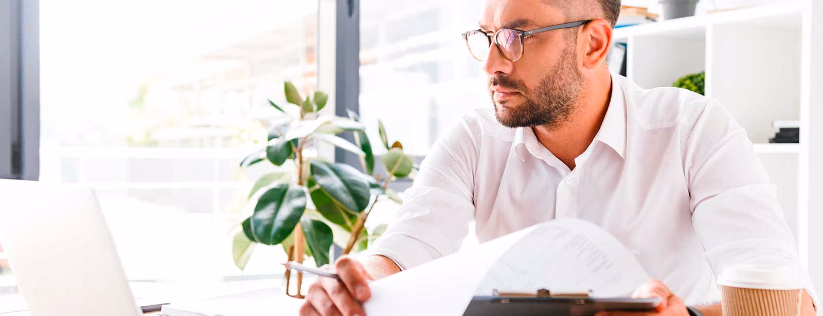 Man in glasses and white shirt reading papers at desk with laptop and coffee cup.